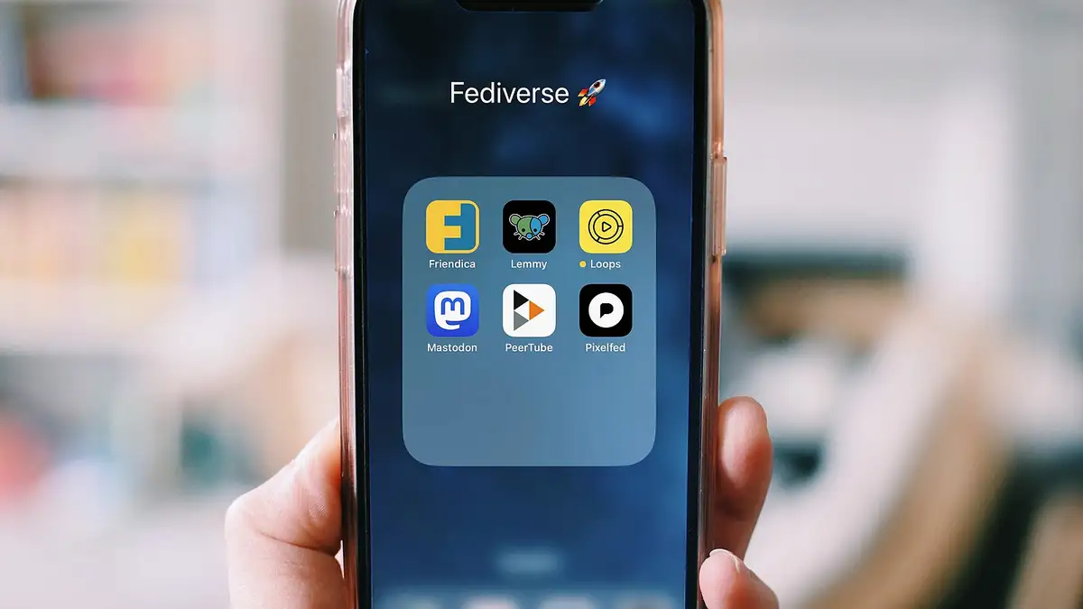a photo of a hand holding a smartphone; on screen you see icons of Fediverse apps: Lemmy, Loops, Mastodon, PeerTube and Pixelfed